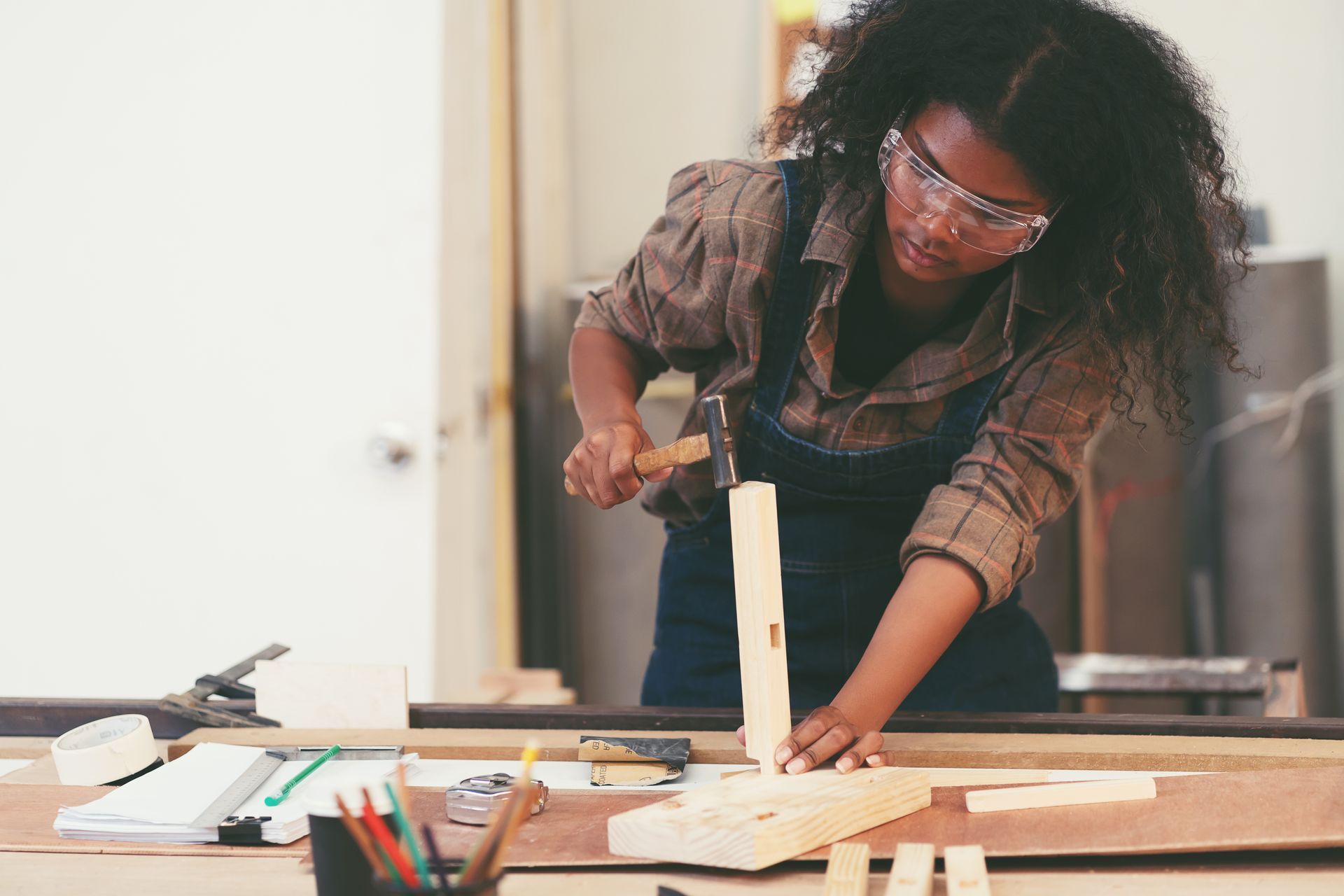 A person wearing safety glasses uses a hammer to join two pieces of wood on a workbench in a workshop.
