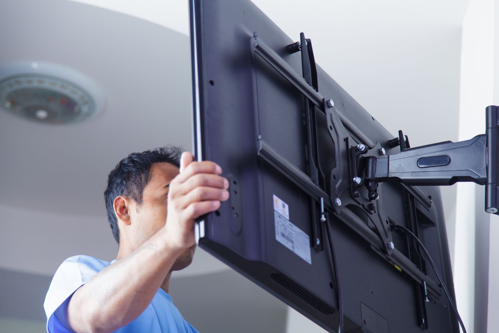 A person in a blue shirt installing a flat-screen television onto a wall-mounted bracket.