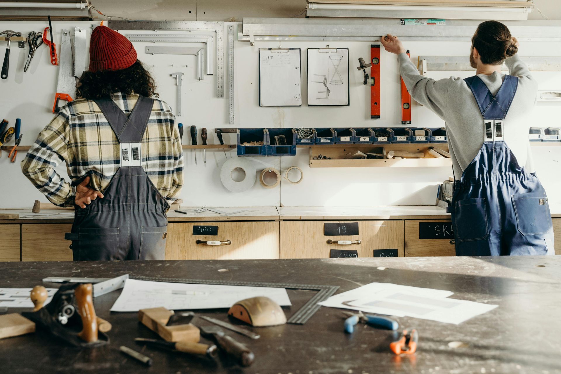 Two people in work overalls stand at a wooden workbench in a workshop, checking tools and plans against a wall display.