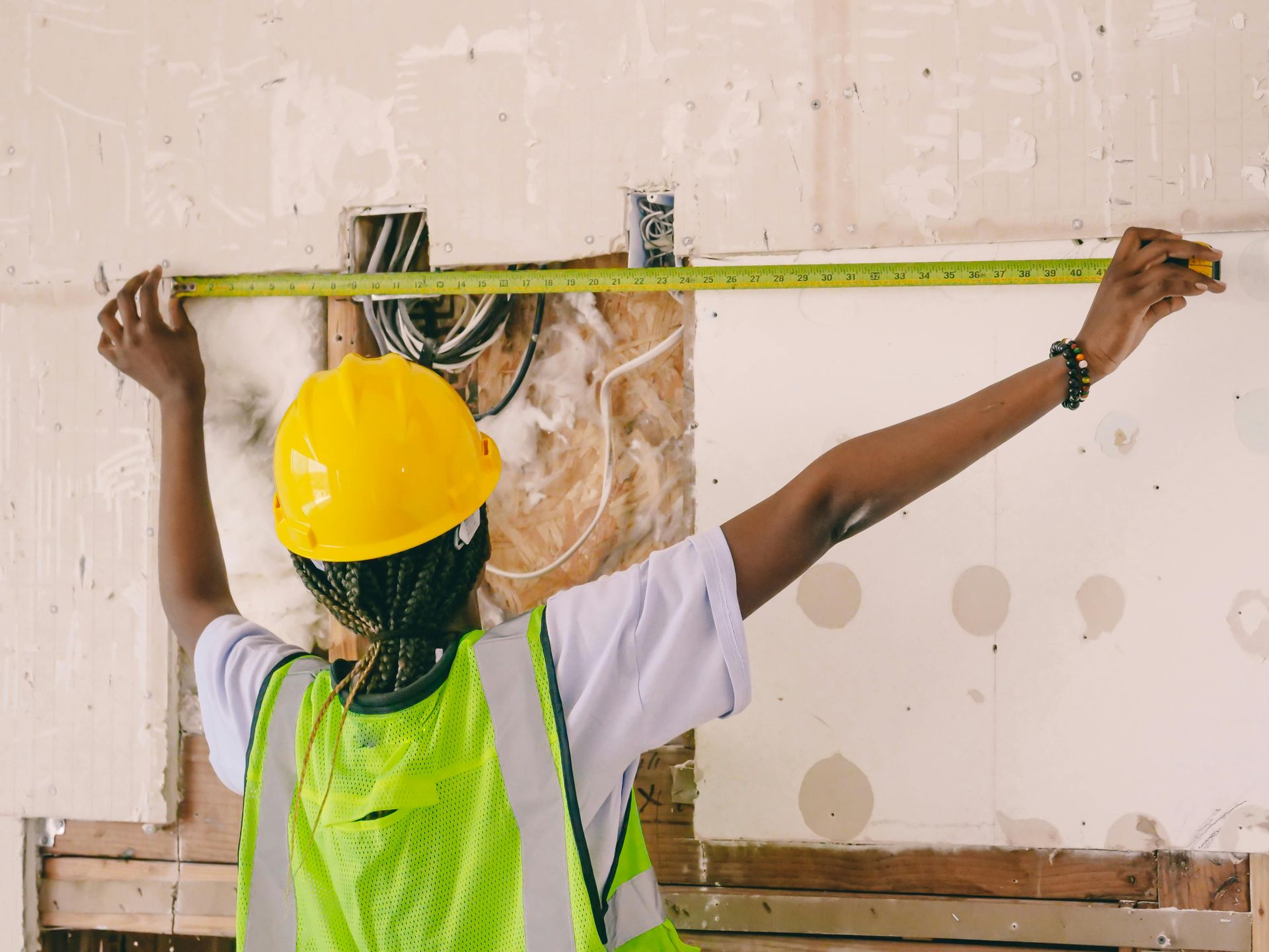 A construction worker in a yellow hard hat and neon vest uses a yellow tape measure to measure a wall under construction.