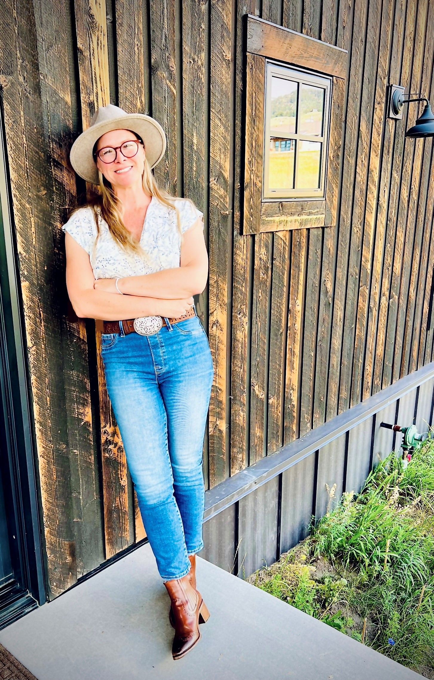 Woman in jeans and a hat leans against a rustic wooden wall, arms crossed.