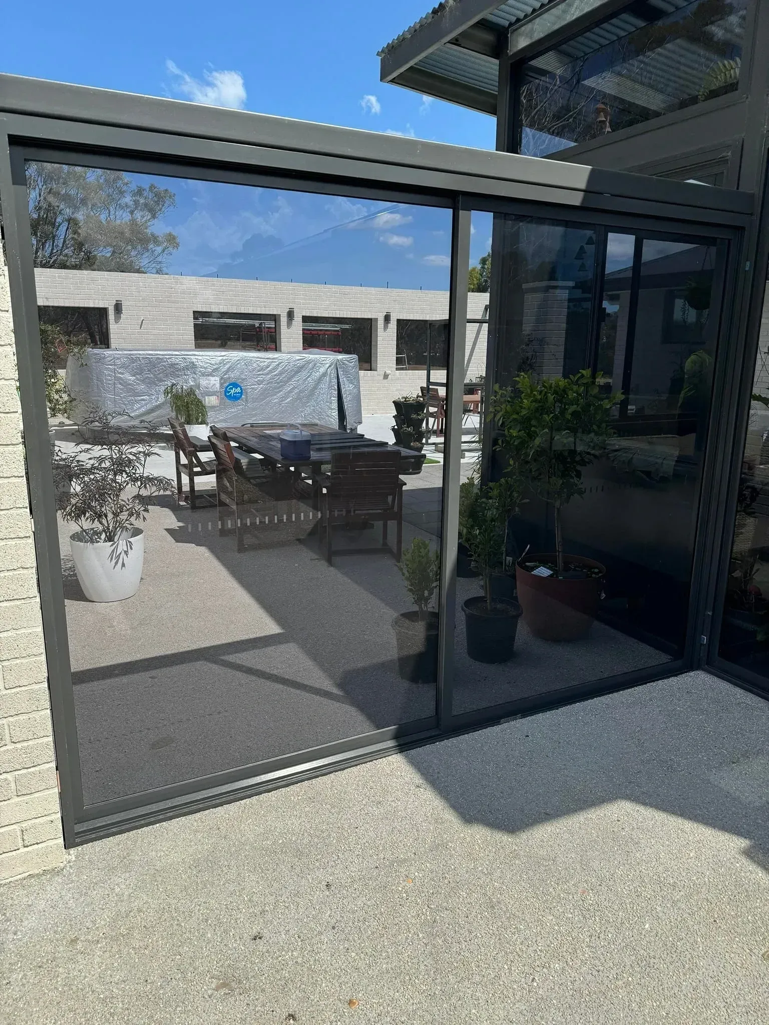 A dark-framed retractable screen door installed on an outdoor patio, showing a blurred view of patio furniture beyond. — Channel Glazing In Electrona, TAS