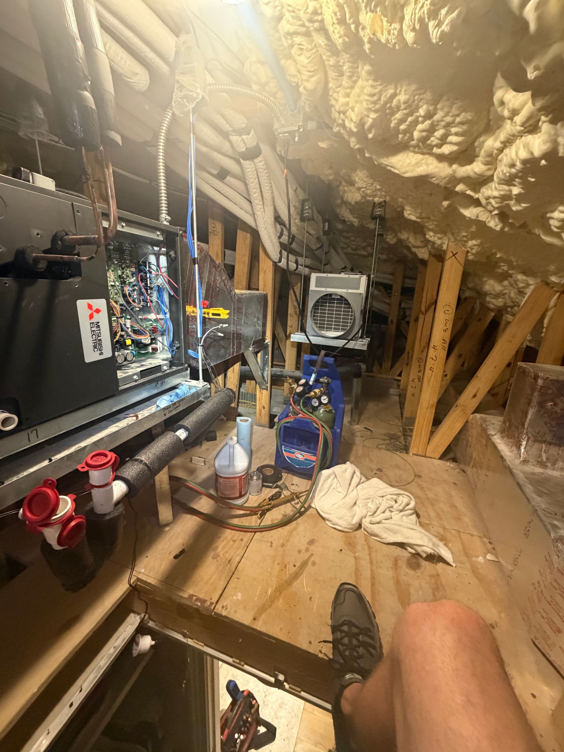 Cluttered attic crawlspace with HVAC equipment, exposed beams, insulation, and a person’s hand in the foreground