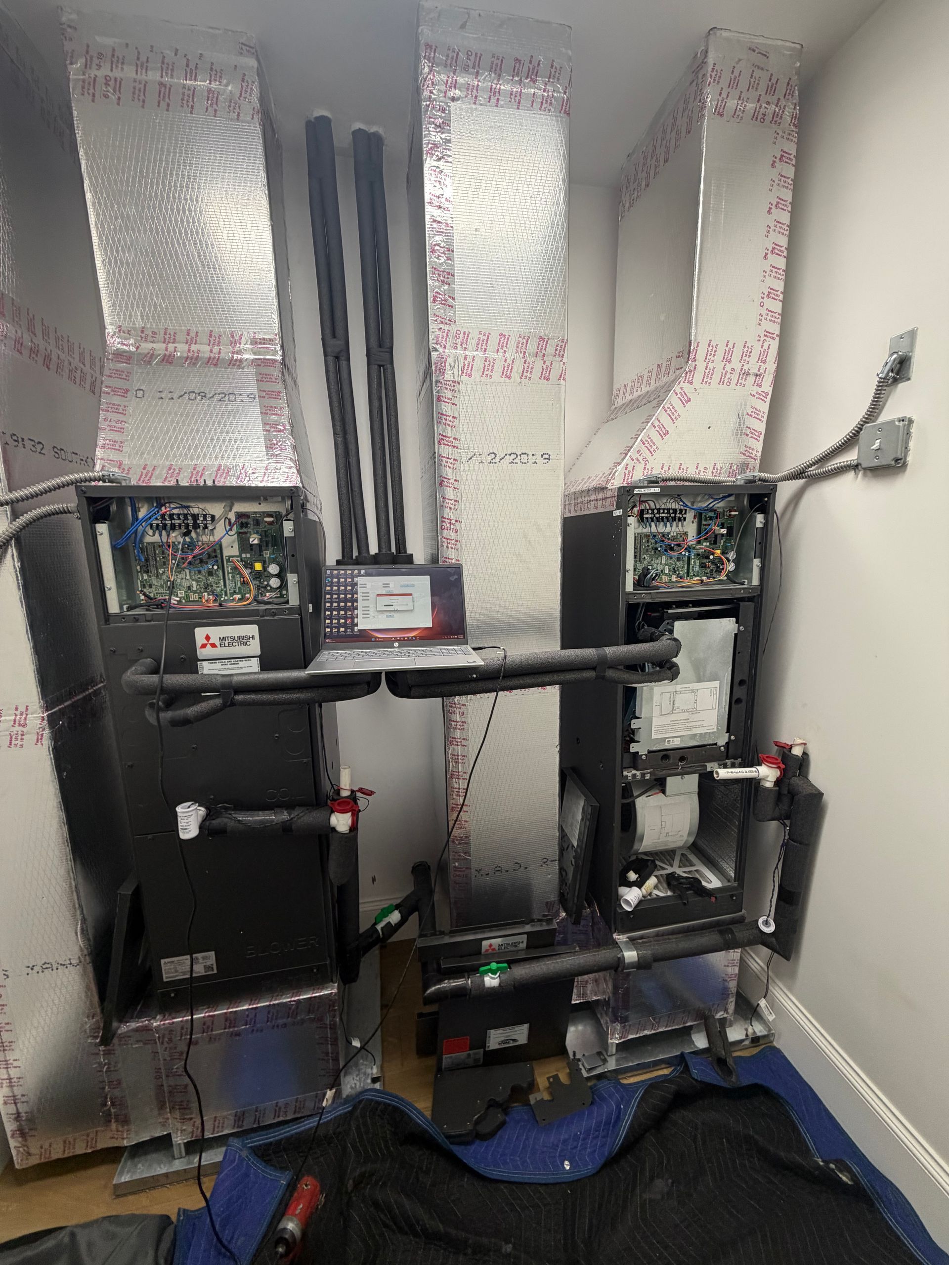 Two black HVAC units in a small utility closet with silver ductwork and blue floor covering.