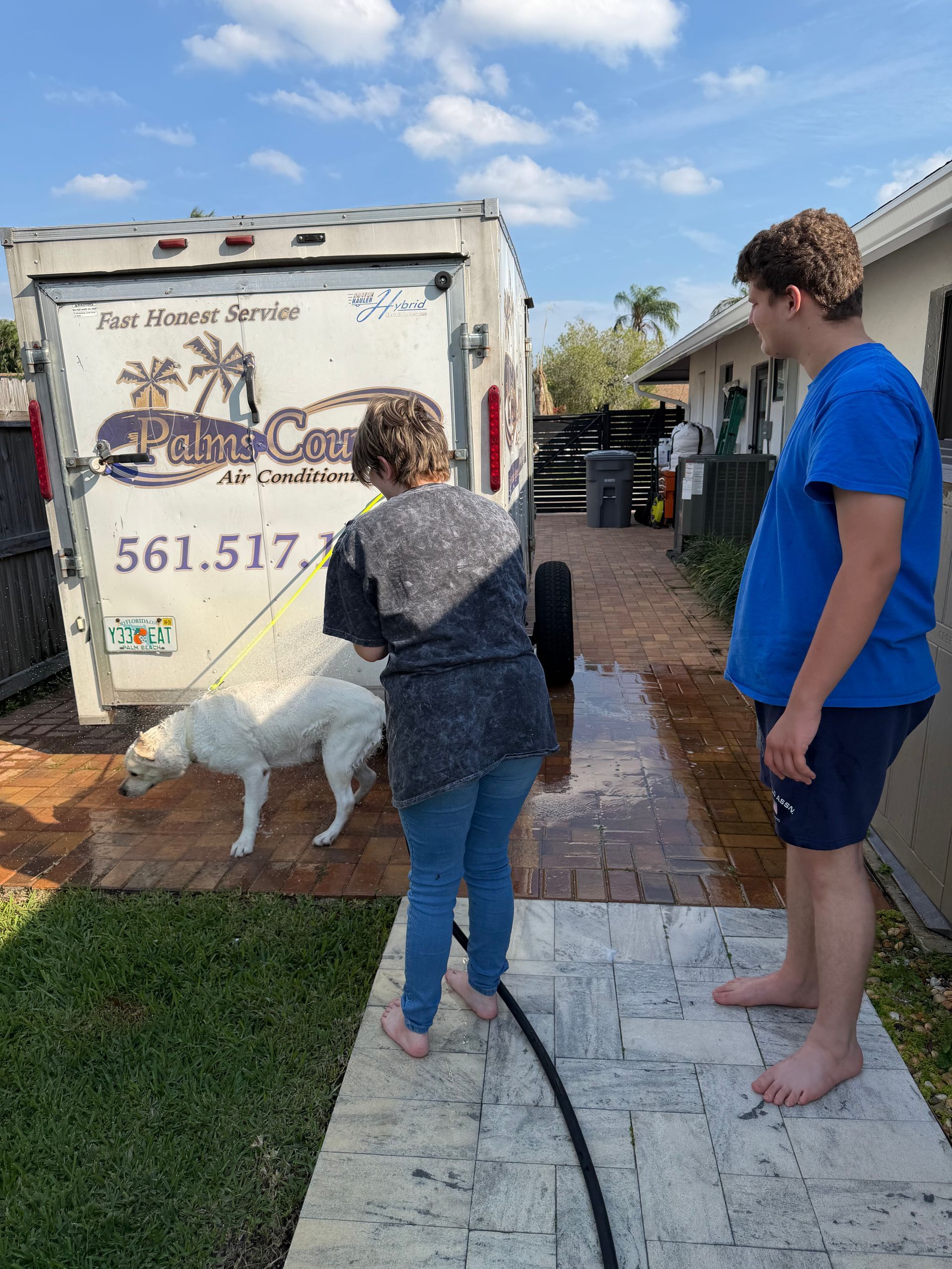 Two people and a white dog outside a small trailer with painted murals and a visible phone number.