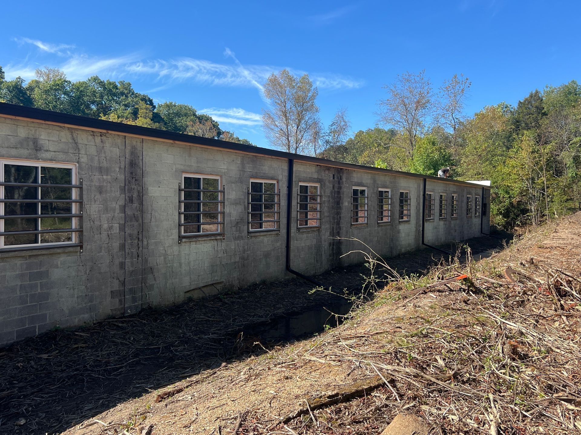 Gray, dilapidated building with barred windows set in a grassy, wooded area under a blue sky.