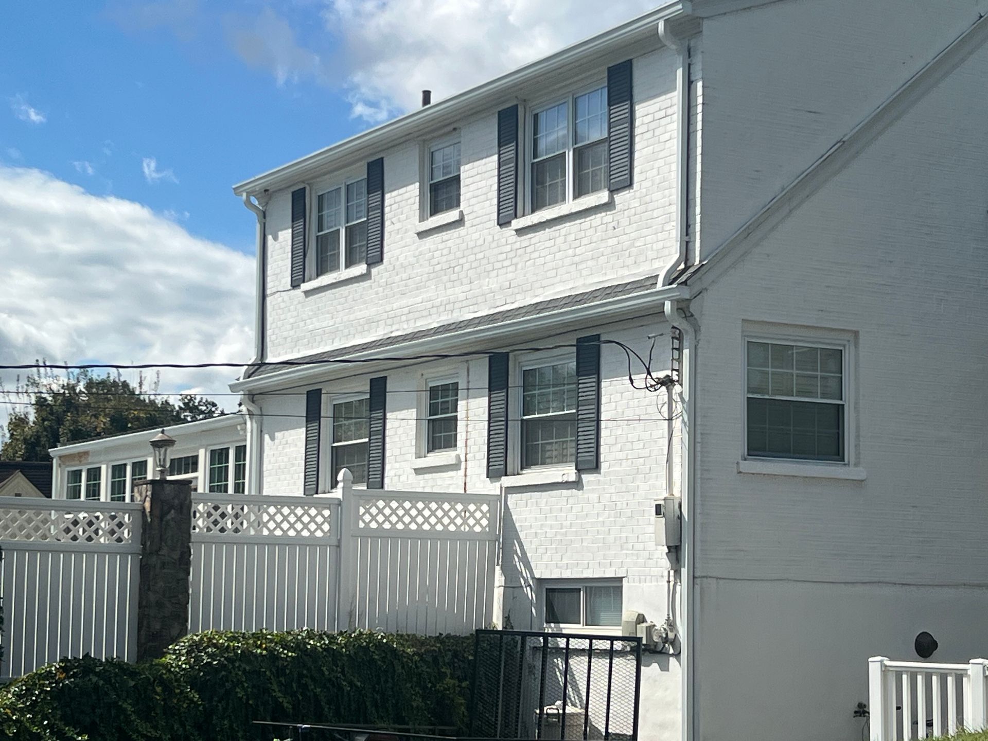 White brick two-story house with black shutters, a white fence, and a blue sky.
