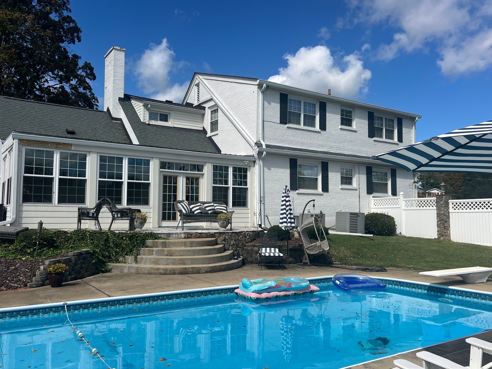 Back of a white two-story house with a pool. Blue sky, black shutters, and a striped umbrella.
