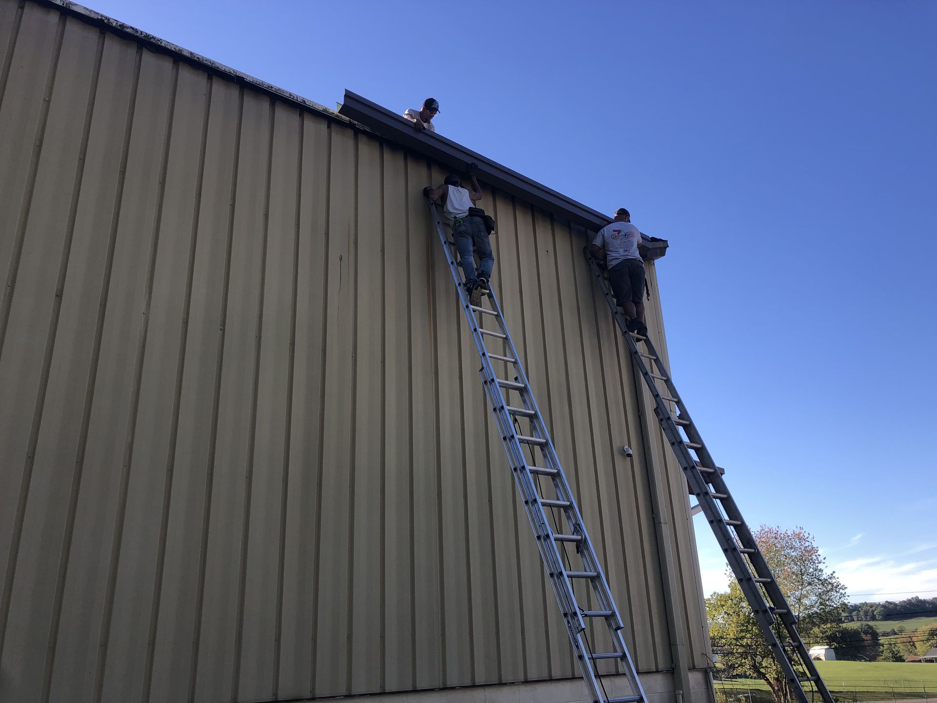 Three workers on ladders installing gutters on a building's exterior under a blue sky.