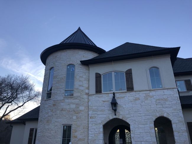 Stone and stucco home with a turret, dark roof, and wooden shutters against a blue sky.