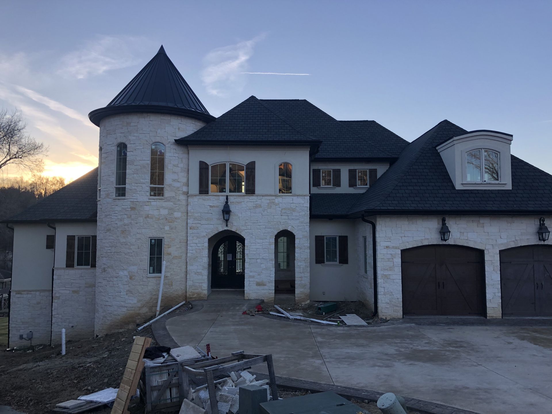 White brick home with black roof, tower, and arched entryway; unfinished driveway.