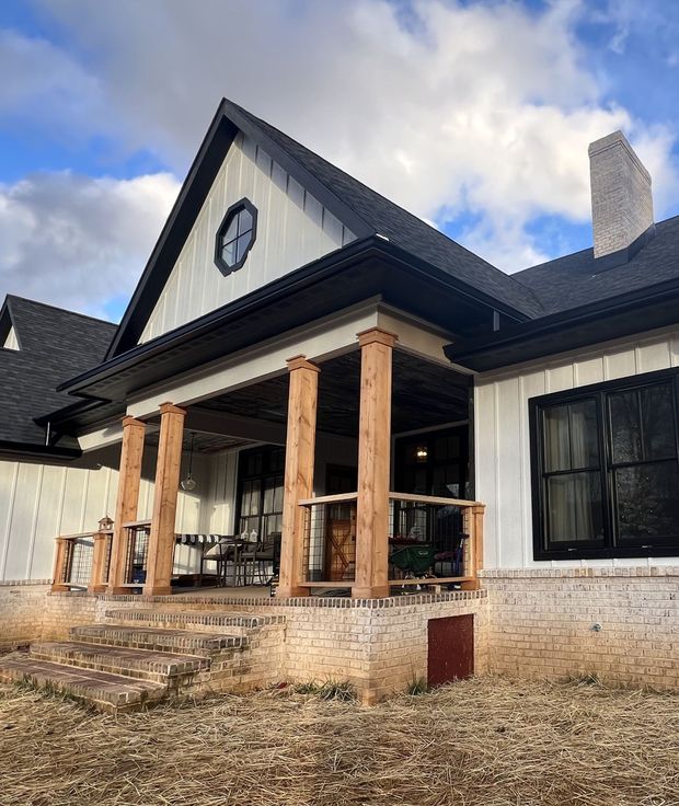 A farmhouse porch with wood columns, white siding, black roof, and a brick foundation.