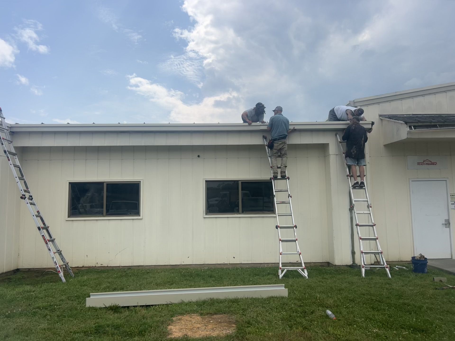 Three people on ladders installing gutter on a white building, under a cloudy sky.