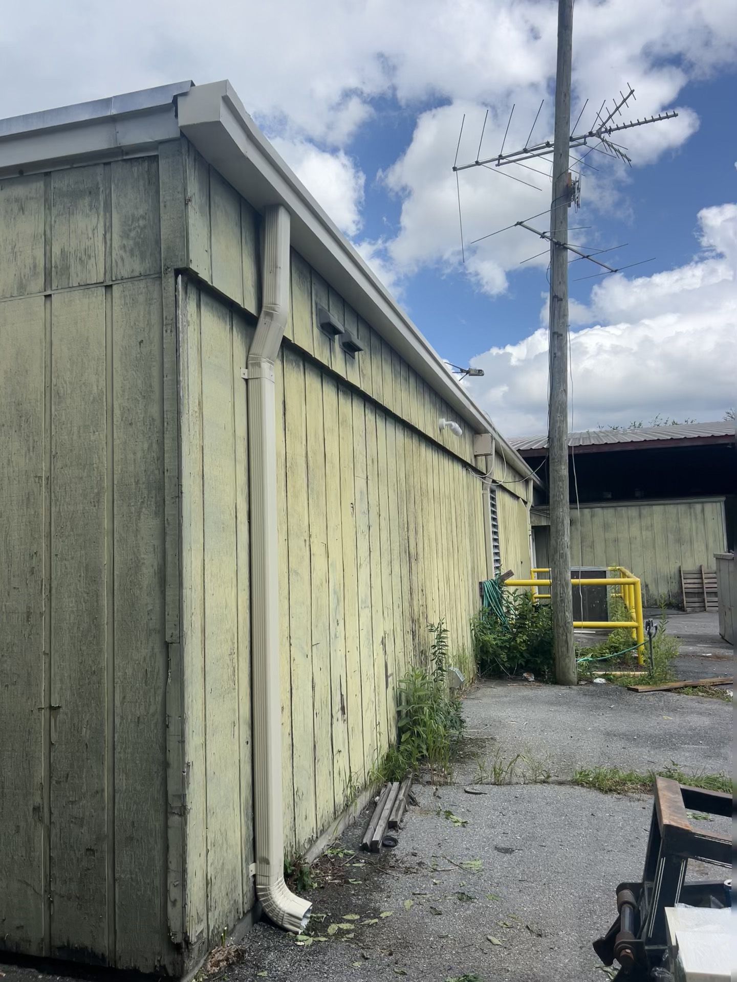 Weathered, light yellow building with white gutters, overgrown vegetation, and a utility pole under a cloudy sky.