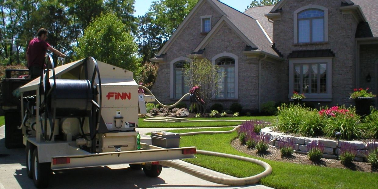 A man operates a machine, spraying mulch onto a lawn of a house with a manicured lawn