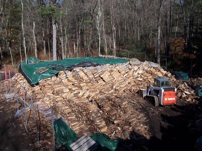 Pile of firewood with a Bobcat tractor, covered by a green tarp, in a wooded area