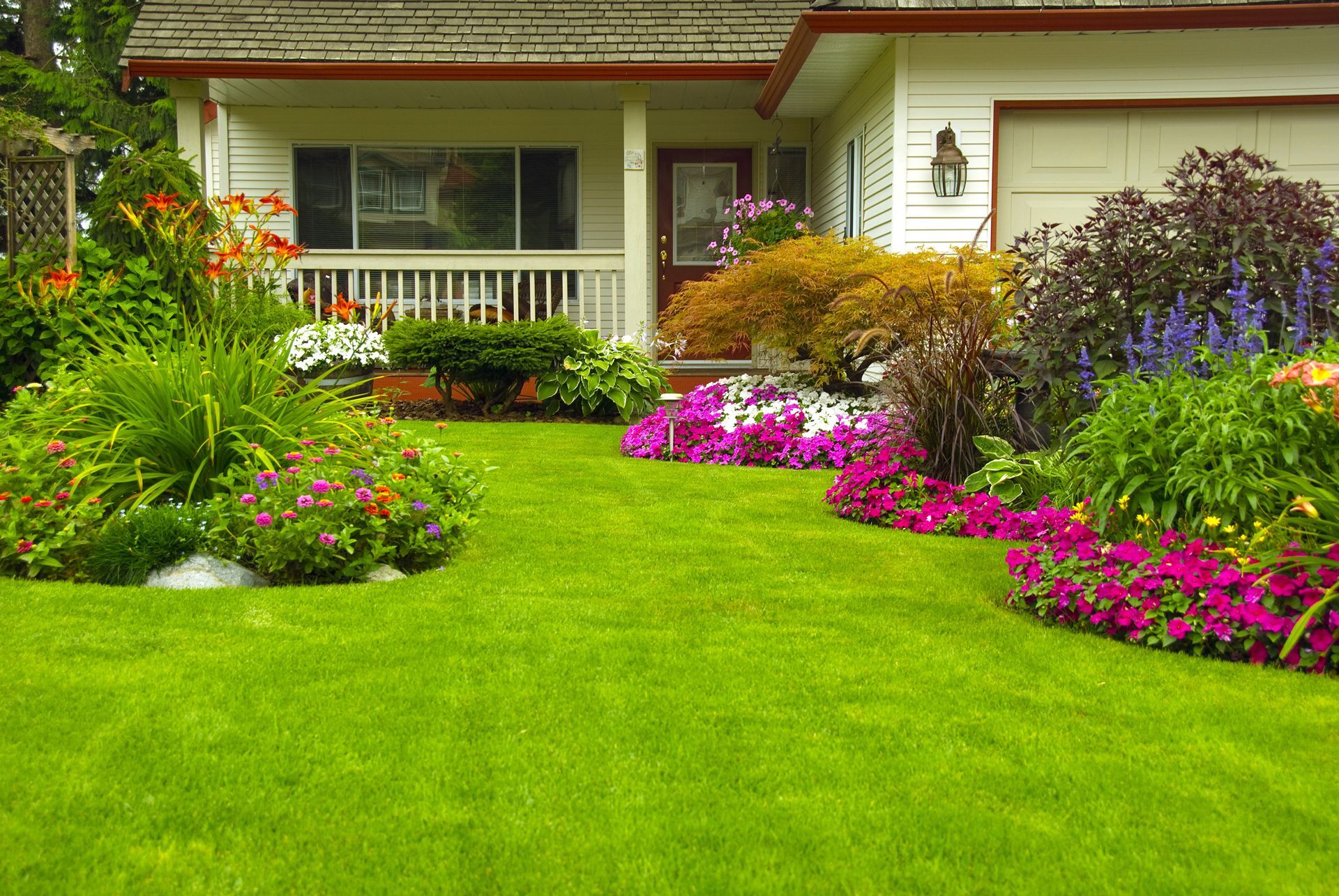Green lawn and colorful flowerbeds in front of a white house with a porch and garage