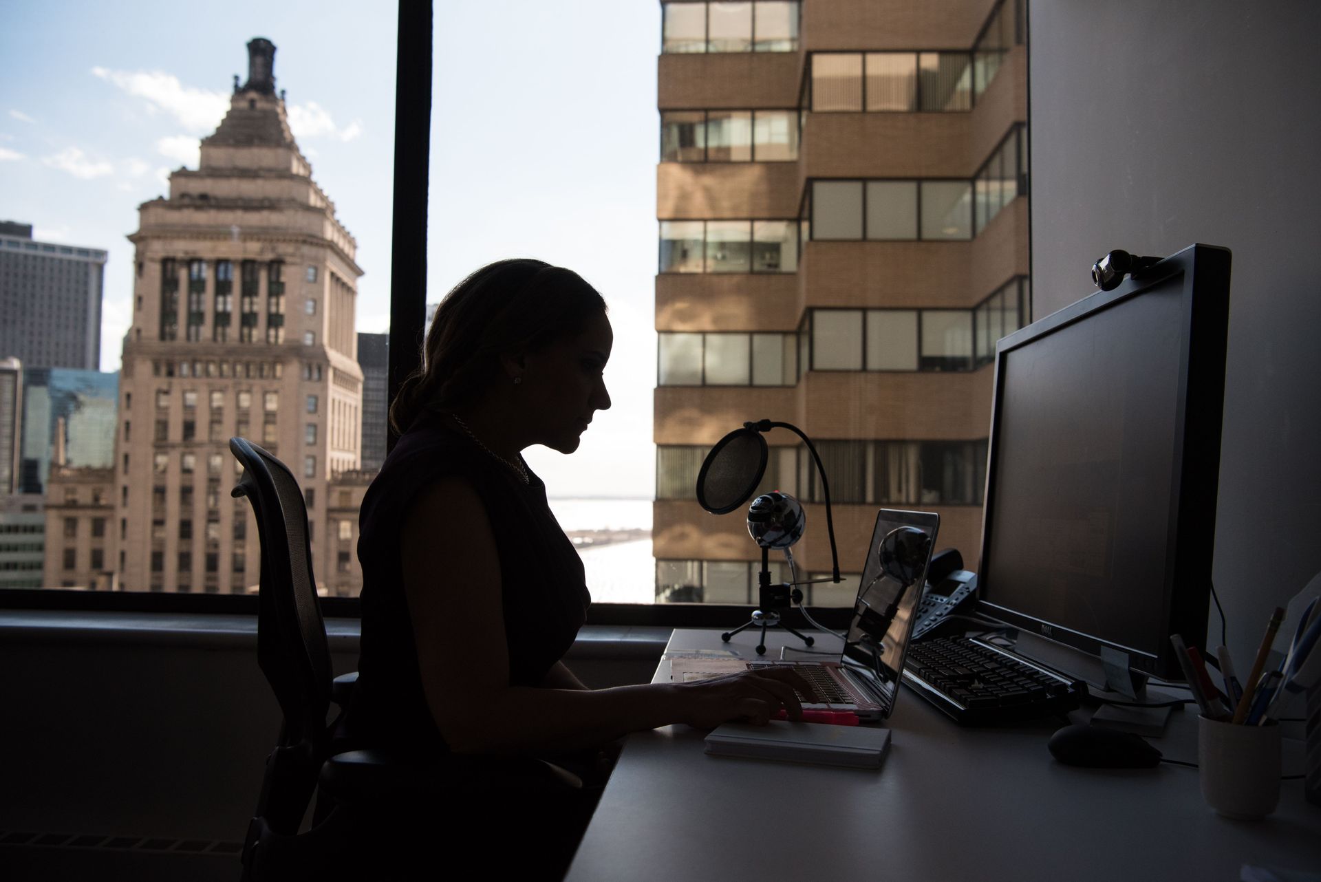 Silhouette of a person working at a desk with a computer and microphone, city buildings visible through a window.