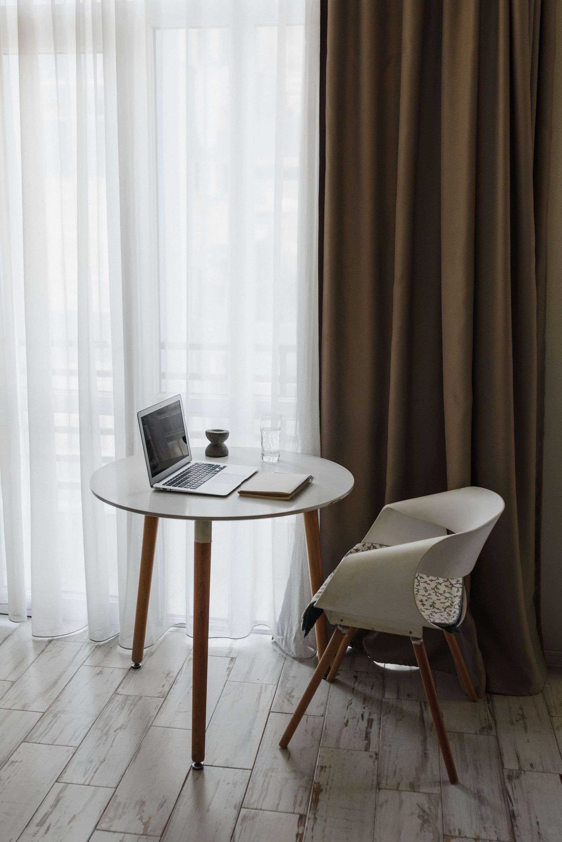 Small round table with laptop and coffee by a chair beside tall curtains in a bright room