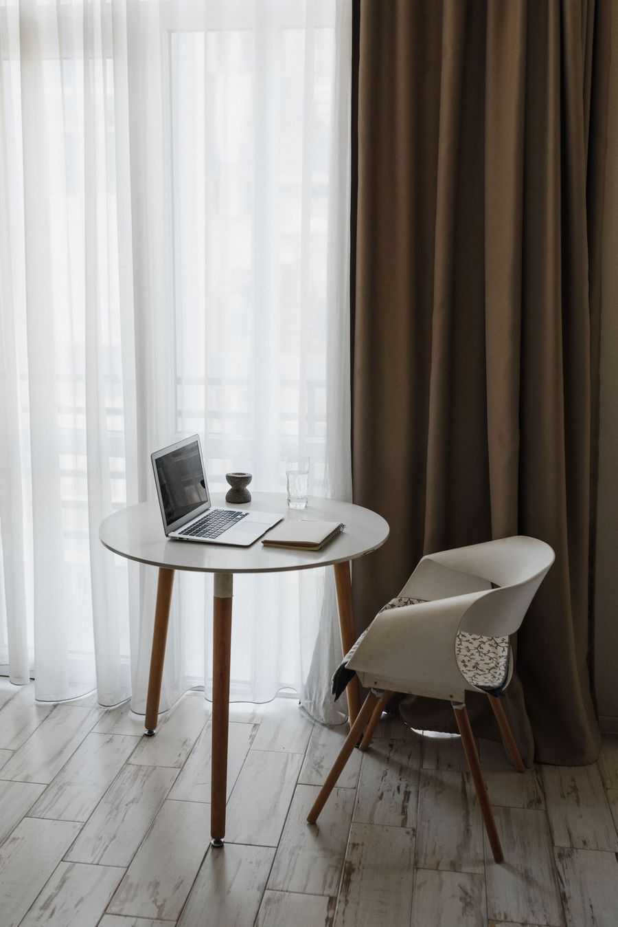 Minimalist workspace with laptop on round table beside a chair, by sheer curtains and dark drapes.