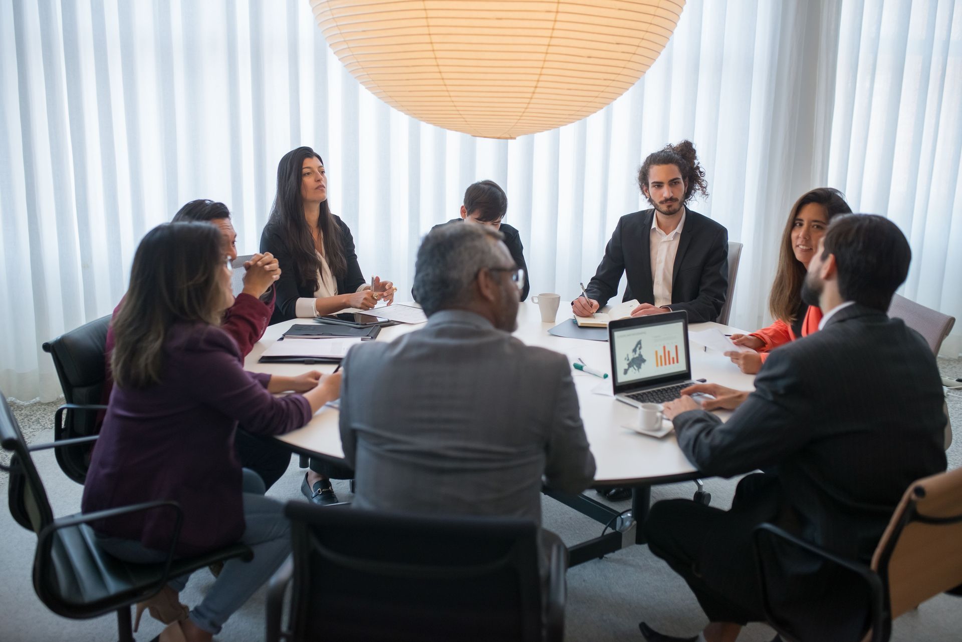 Meeting room with six people seated around a table, discussing a presentation on a laptop screen