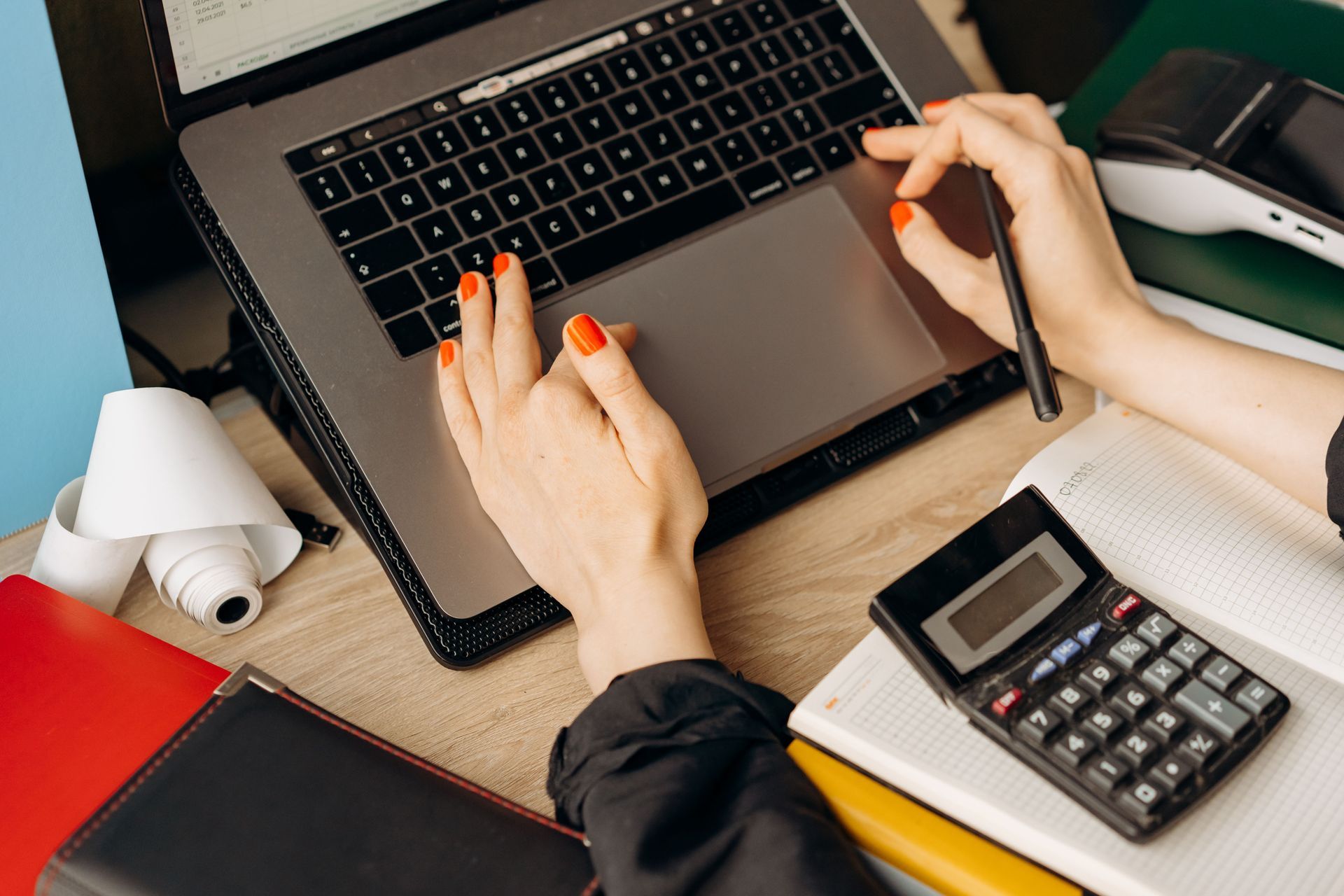 Hands typing on a laptop beside a calculator, notebook, and red folder on a desk
