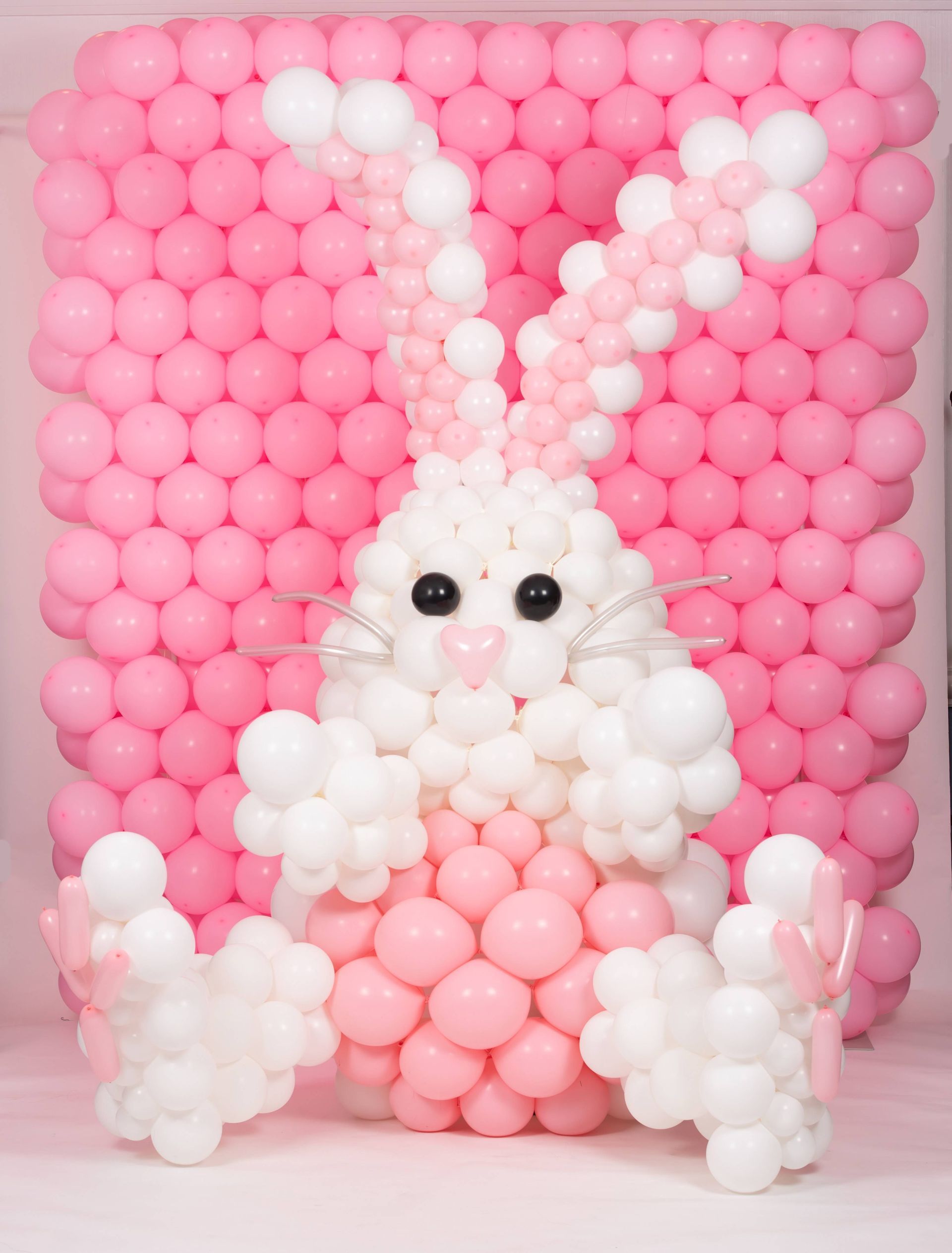 A bunny made out of pink and white balloons is sitting in front of a wall of pink balloons.