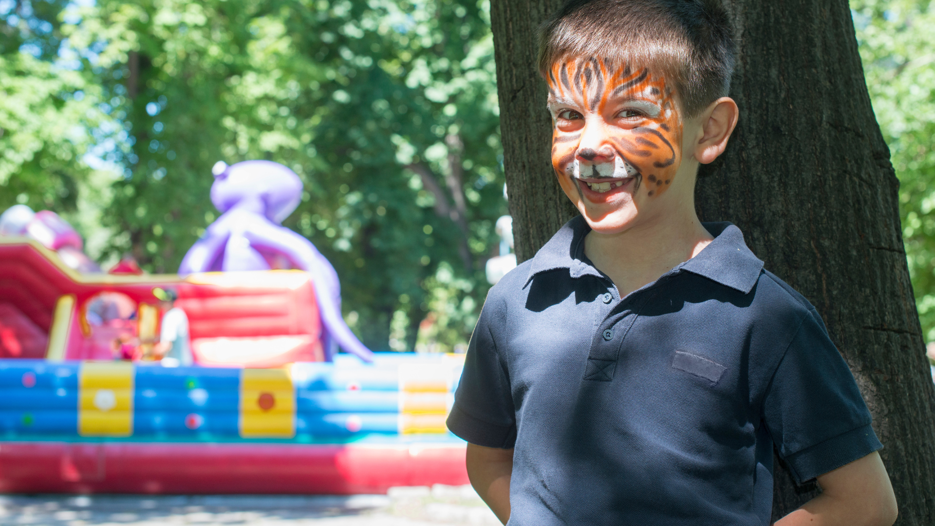 A young boy with his face painted like a tiger is standing next to a tree.