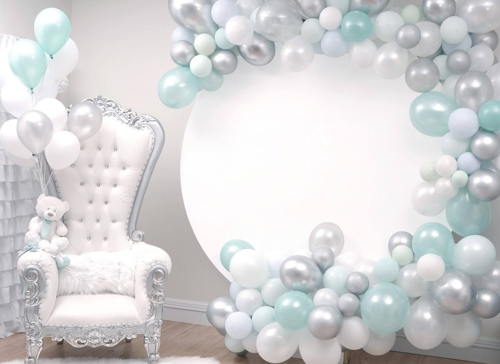 A white chair with a teddy bear sitting on it is surrounded by balloons.
