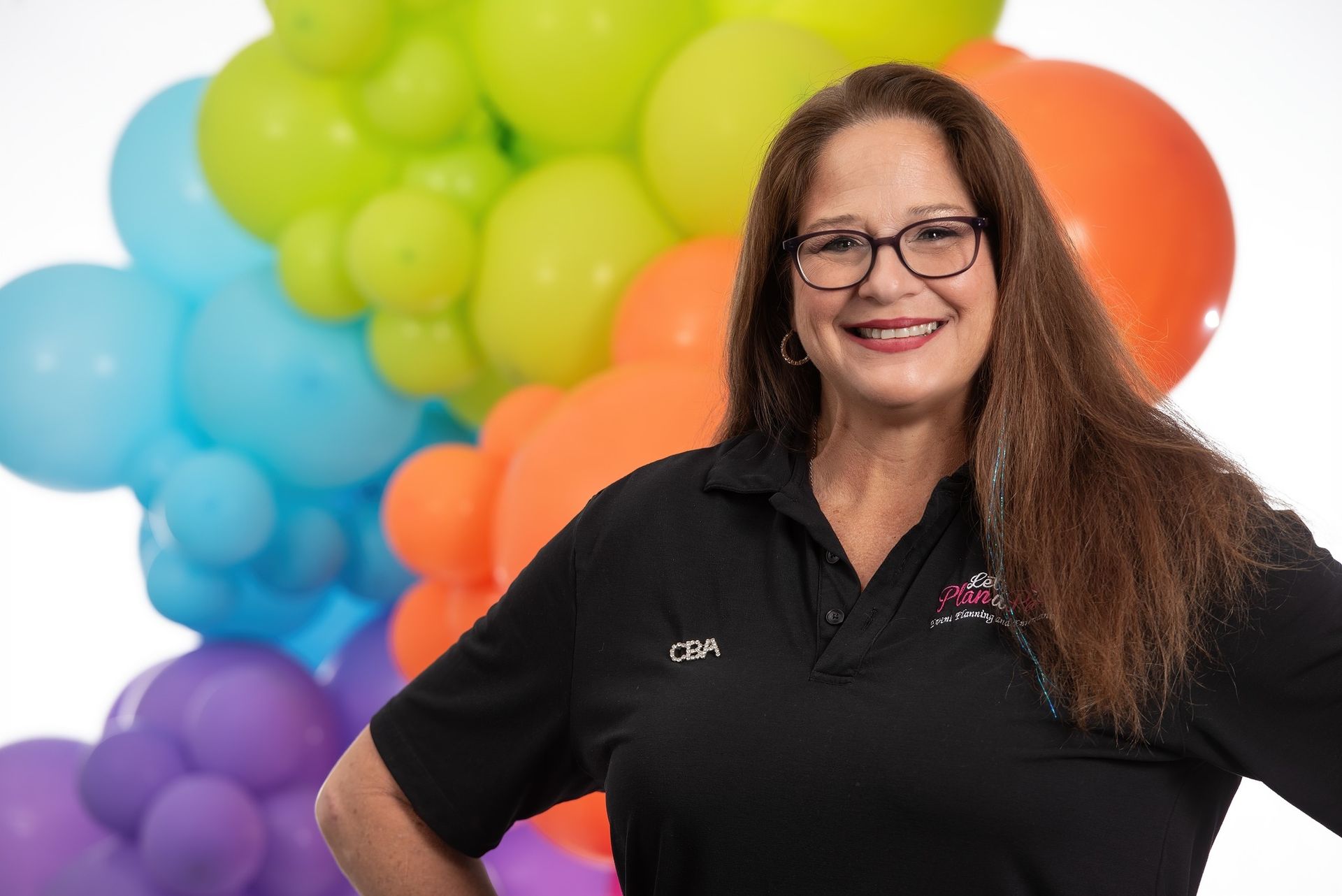 A woman wearing glasses and a black shirt is standing in front of a bunch of colorful balloons.