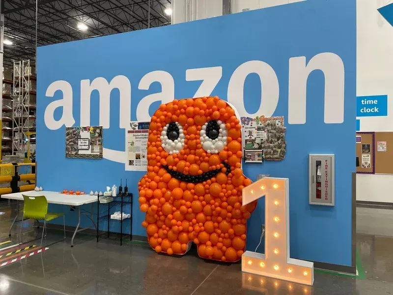 A mascot made out of orange balloons is standing in front of an amazon sign.