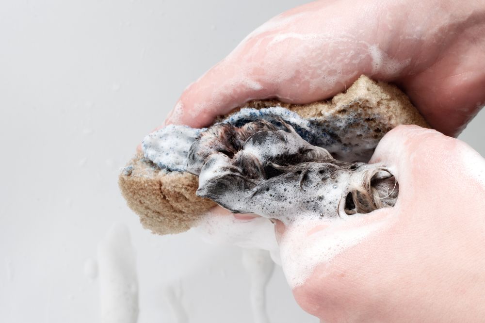 Hands squeezing a soapy sponge over a white surface, with a dark, wet cloth inside.