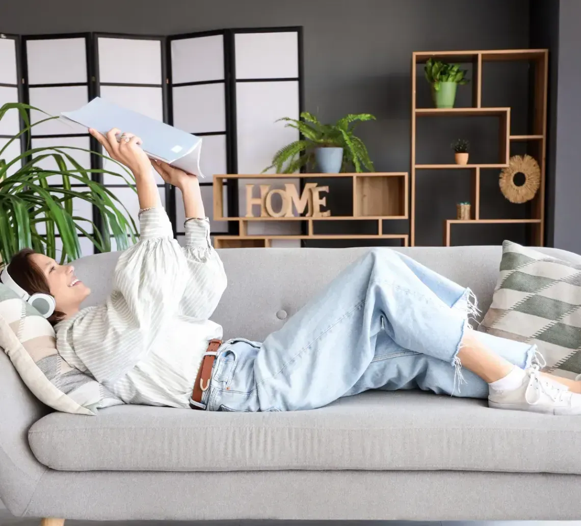 A person wearing headphones lies on a sofa, holding a laptop up in the air while smiling in a brightly lit living room.