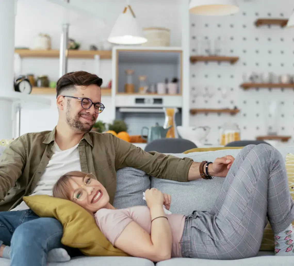 A couple sits together on a grey sofa in a brightly lit, modern living room, smiling and relaxing.