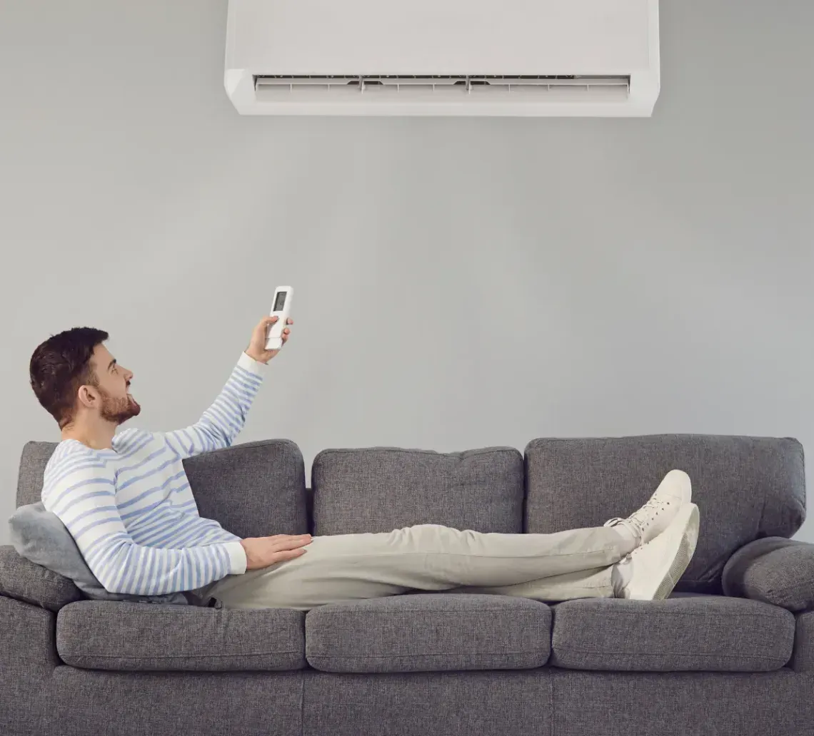 A person relaxes on a gray sofa while aiming a remote at an air conditioner mounted on a gray wall.