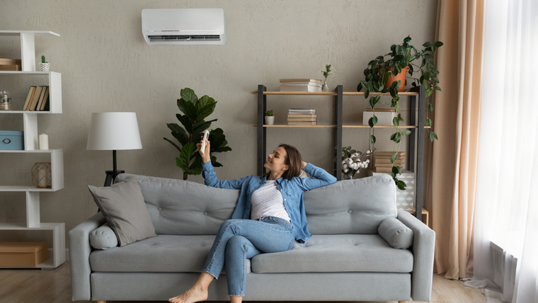 A person sitting on a sofa in a living room uses a remote to adjust a wall-mounted air conditioner.