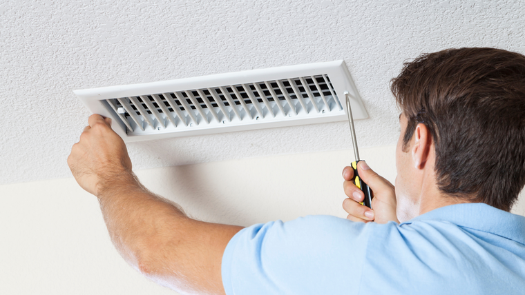 A technician in a blue shirt uses a drill to secure a white air vent to a textured white ceiling.