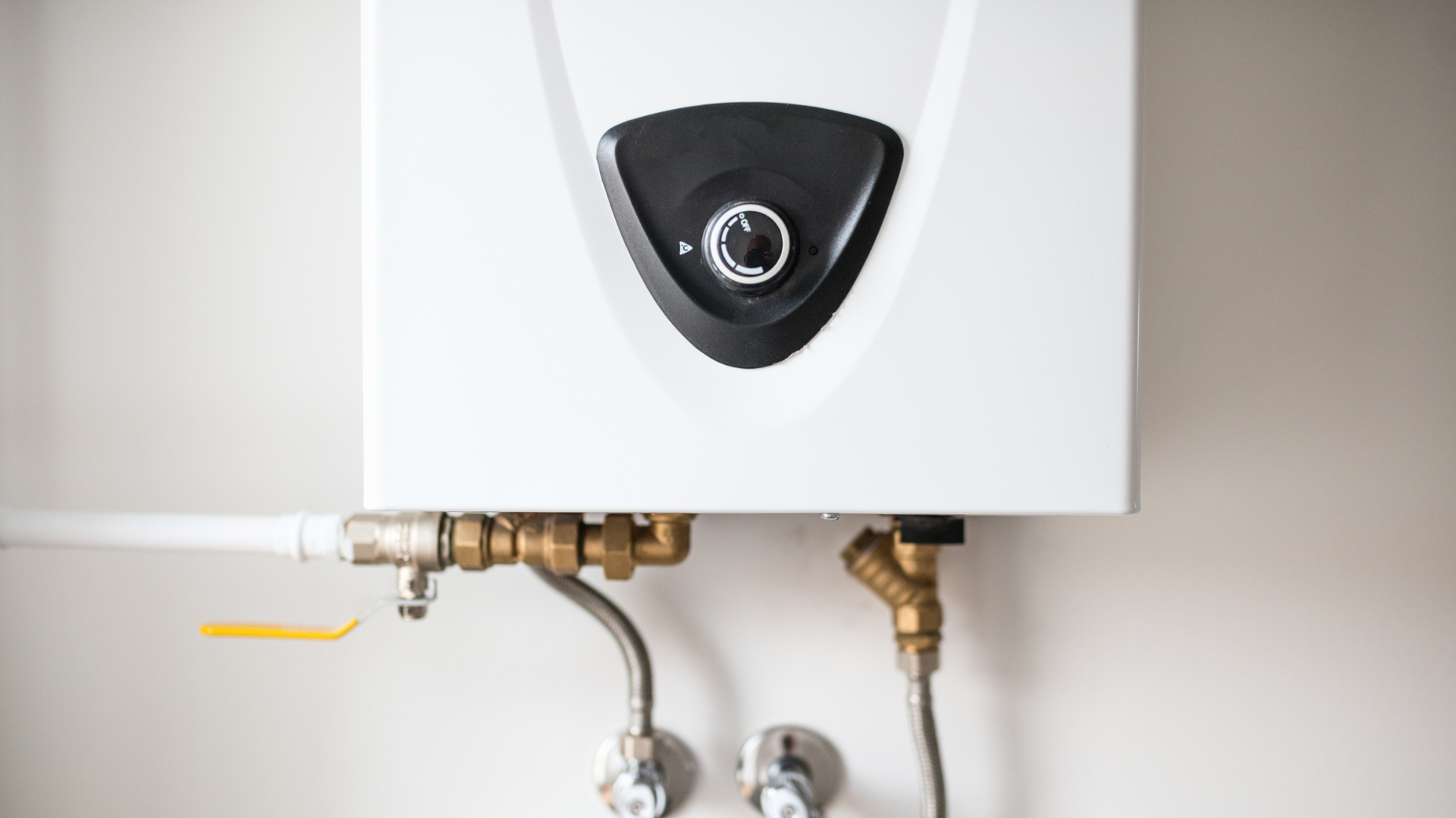 A white electric tankless water heater mounted on a wall with metal plumbing pipes and valves connected at the bottom.