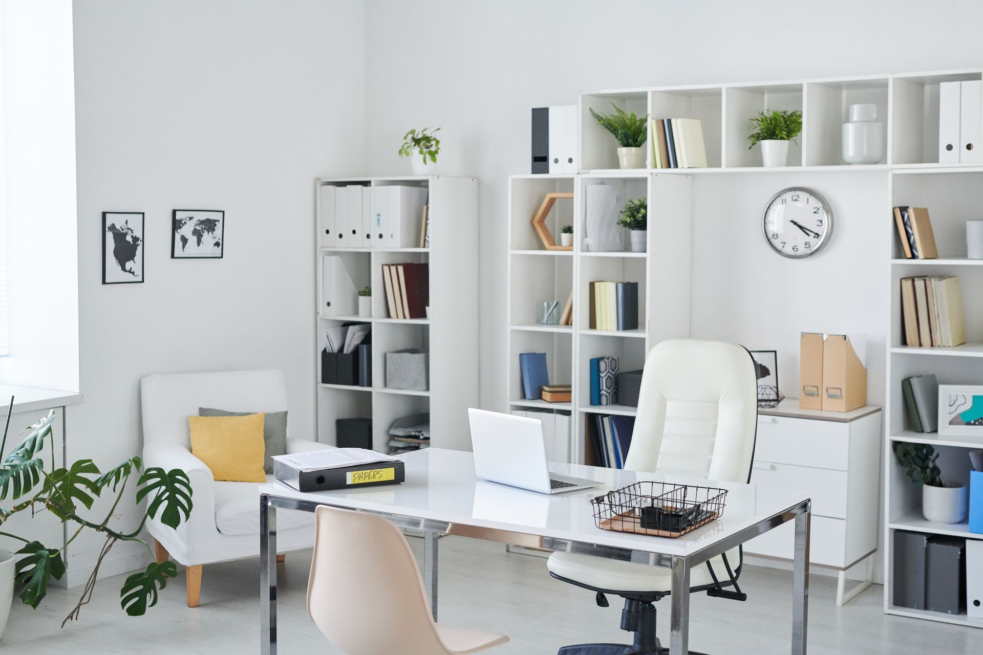 Bright white office with a desk, chair, and shelves filled with books, plants, and files. Includes a clock, artwork, and a laptop.