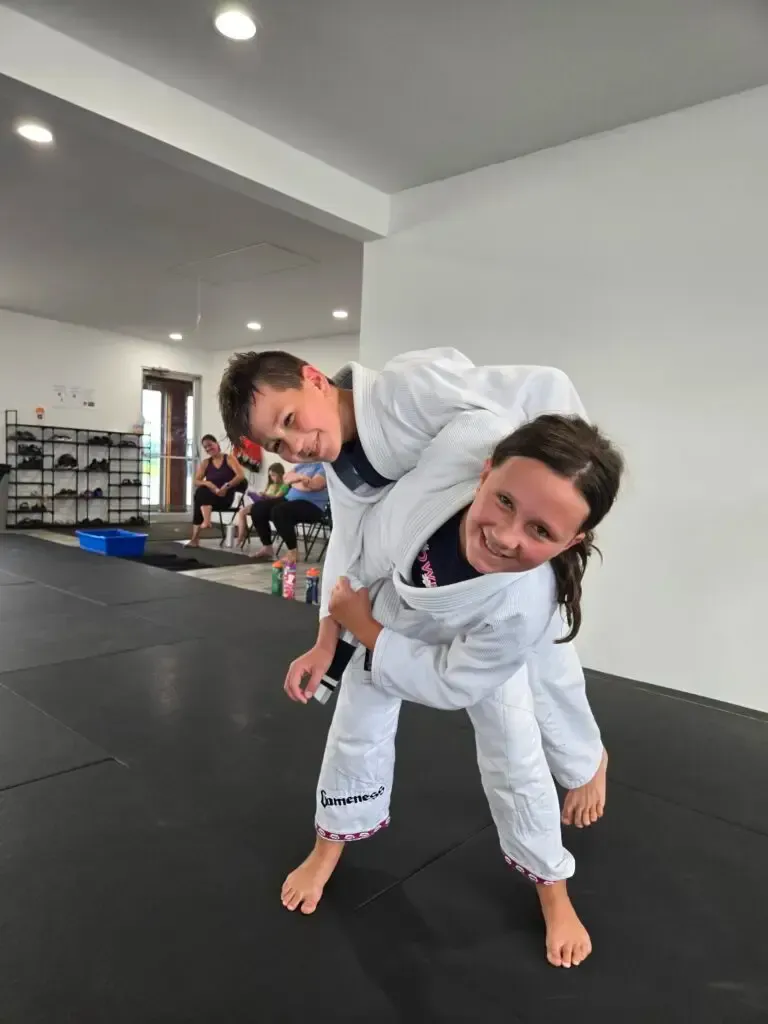 Two children in white martial arts uniforms practice a takedown on a black mat in a gym.