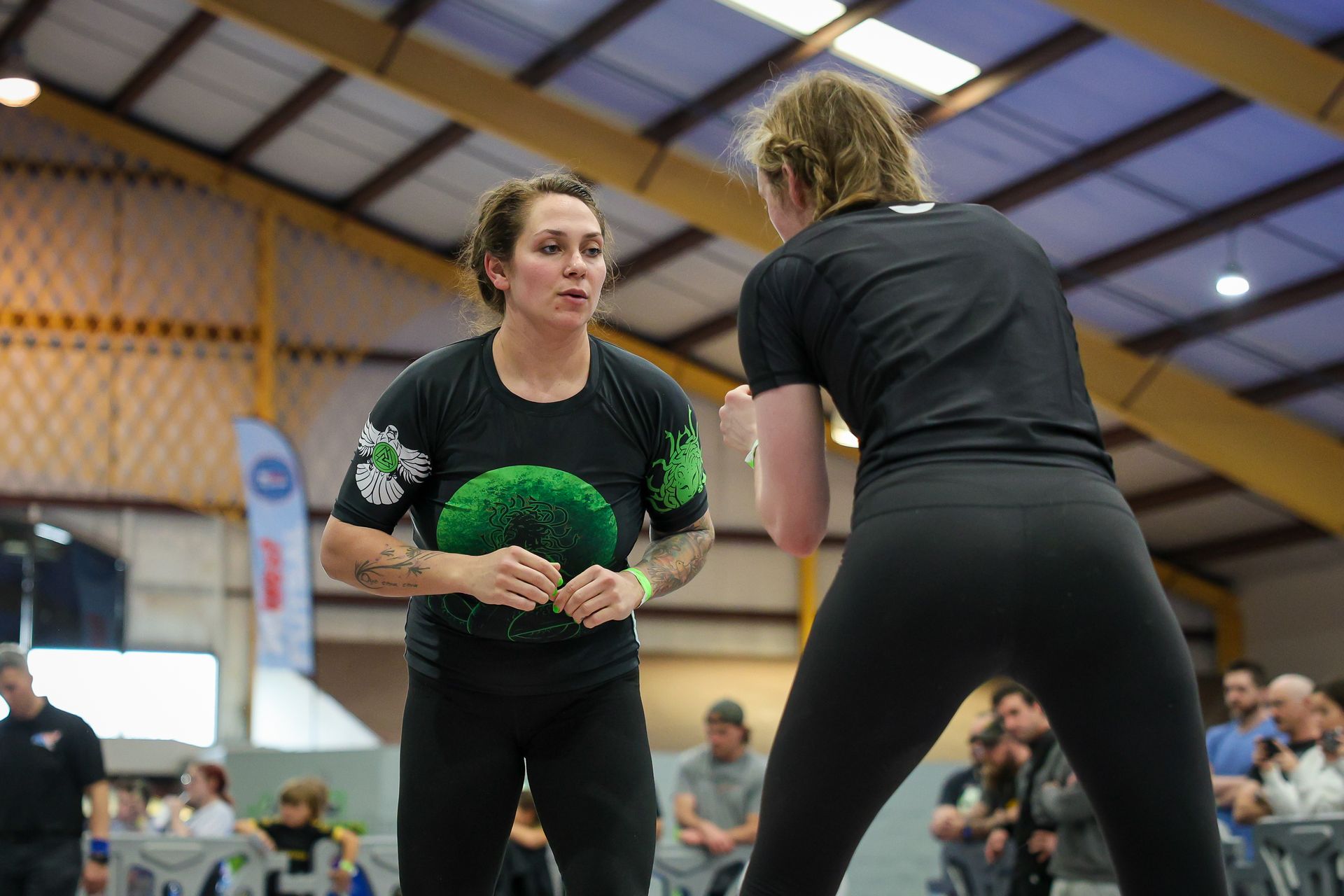 Two women in black athletic wear face each other in a gymnasium, possibly sparring.