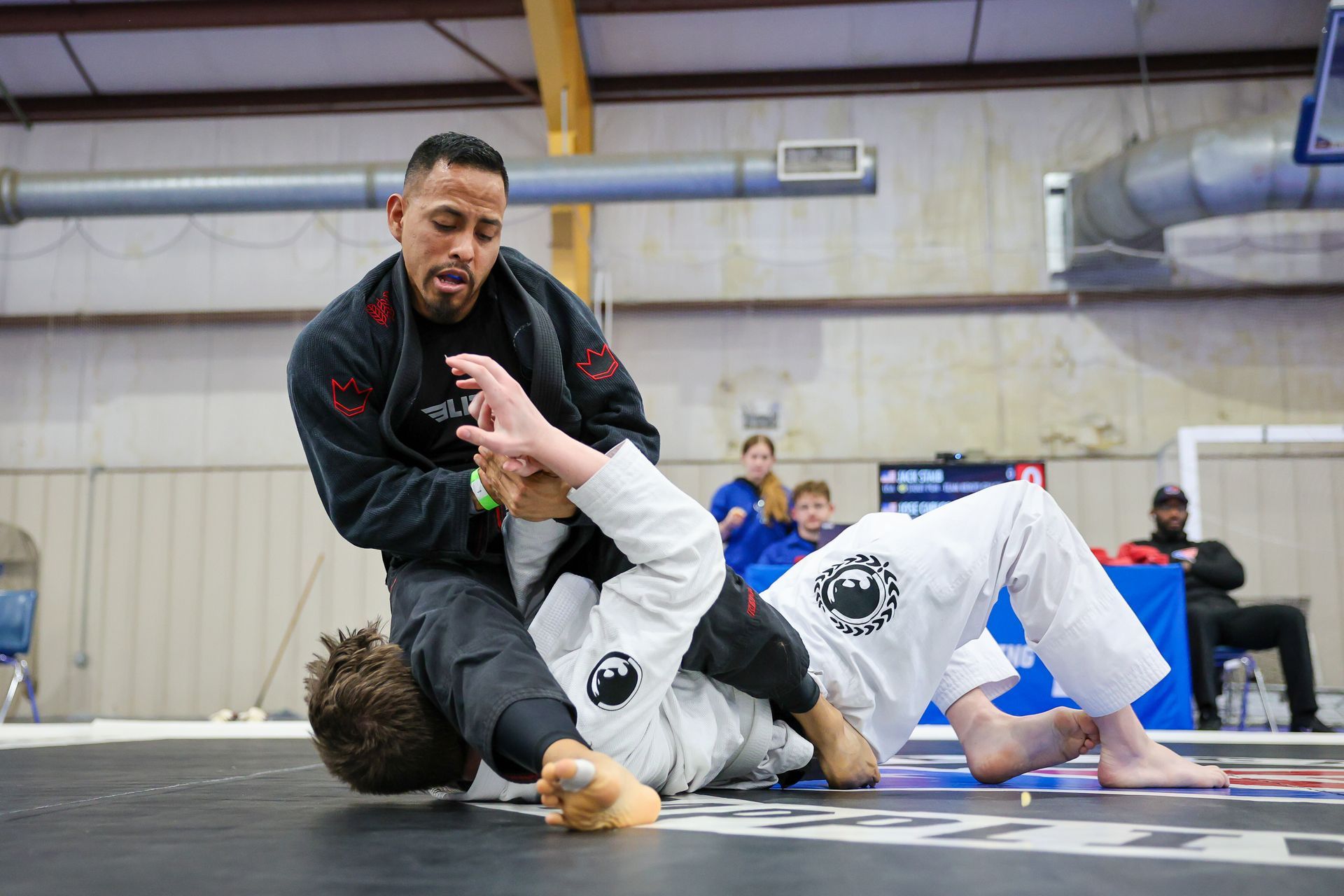 Two men in a grappling match, one on top in dark gi, securing a hold on the other, white gi, on the ground. Indoors.