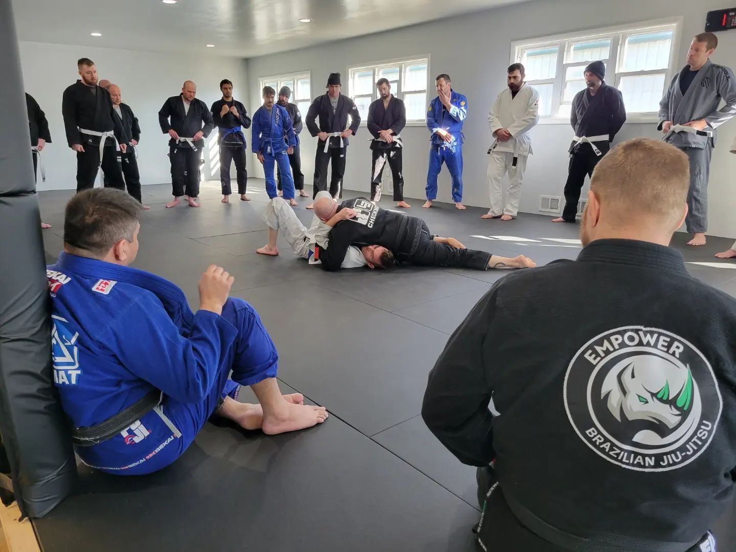 Brazilian Jiu-Jitsu class. Students in gis watch two practitioners grappling on a mat. Interior setting, bright lighting.