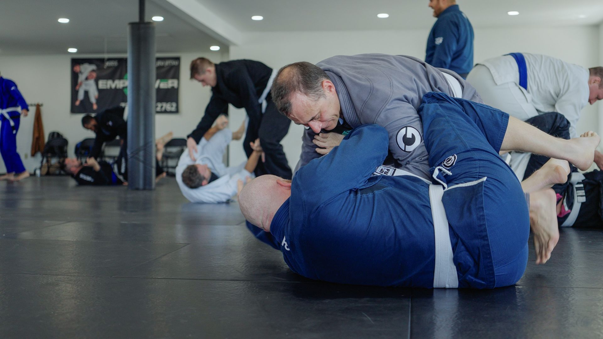People practicing Jiu-Jitsu on a black mat in a gym. One person is on top of another in a grappling position.