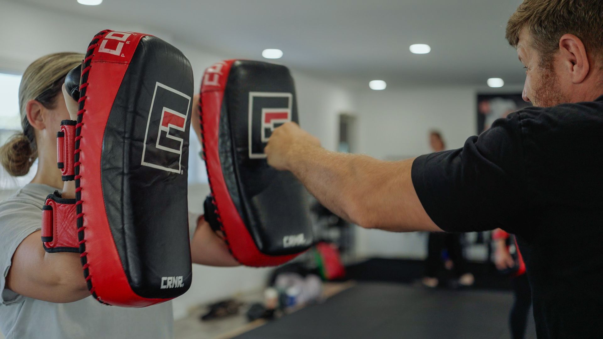 Man holding kick pads for woman during martial arts training in a gym.