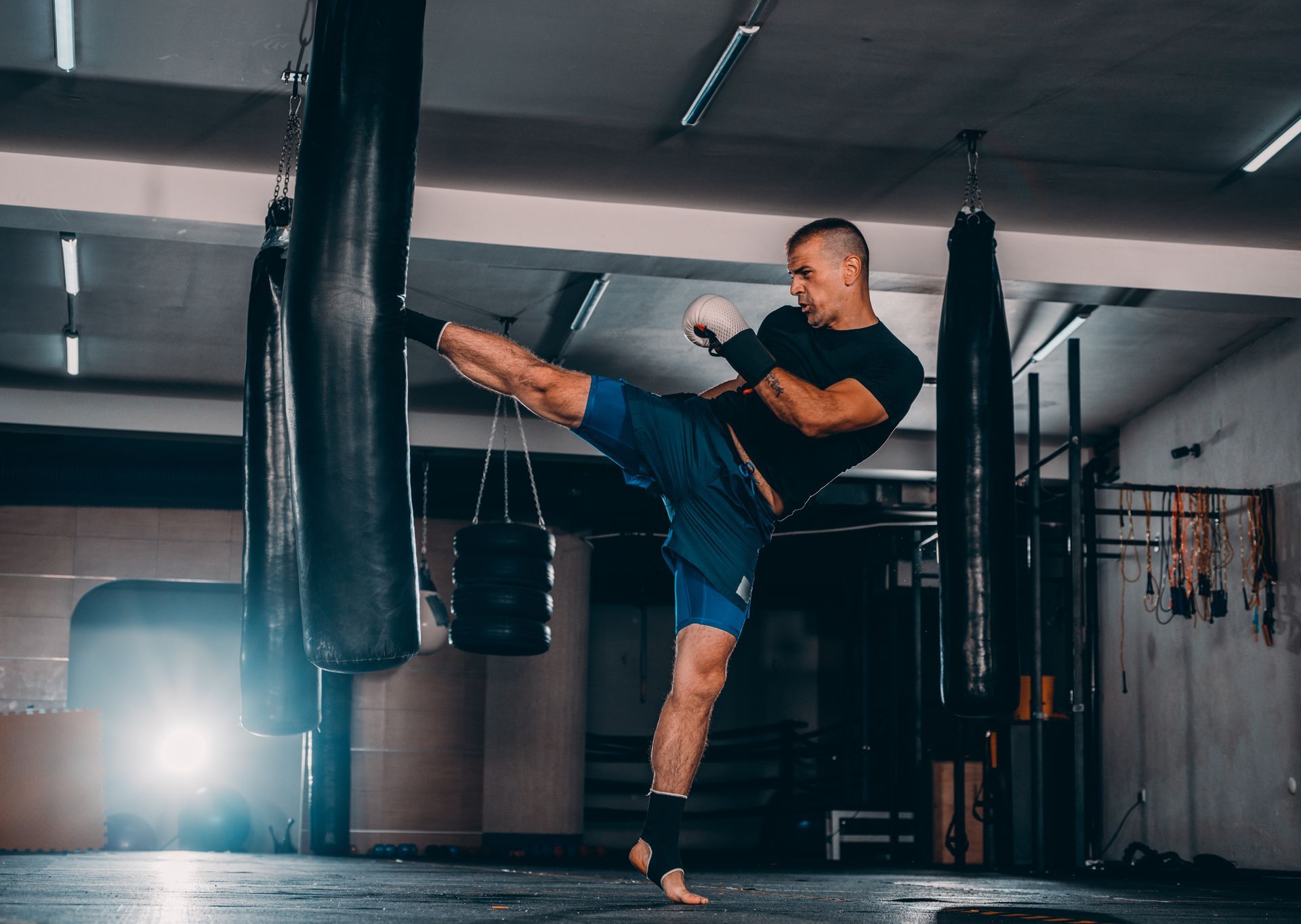 Man kicking a heavy bag in a gym. He wears gloves, shorts, and ankle supports.