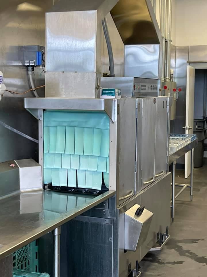 Commercial dishwasher in a stainless steel kitchen. Blue curtains hang from the entry.