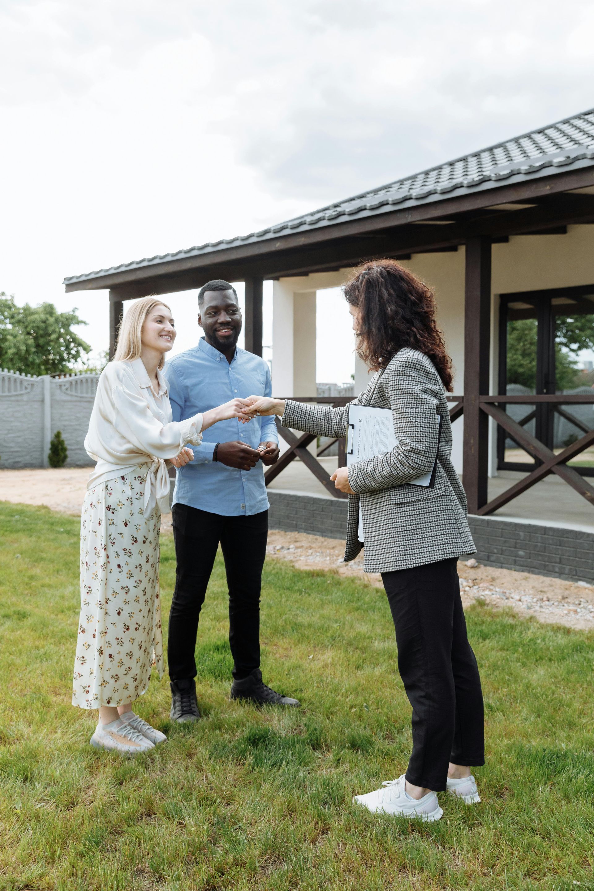 Three people stand on grass shaking hands outside a house with a tiled roof.