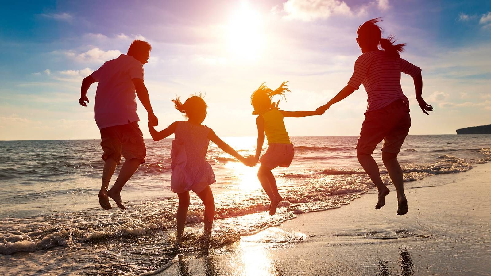 Family of four holding hands, jumping on a beach with the setting sun.