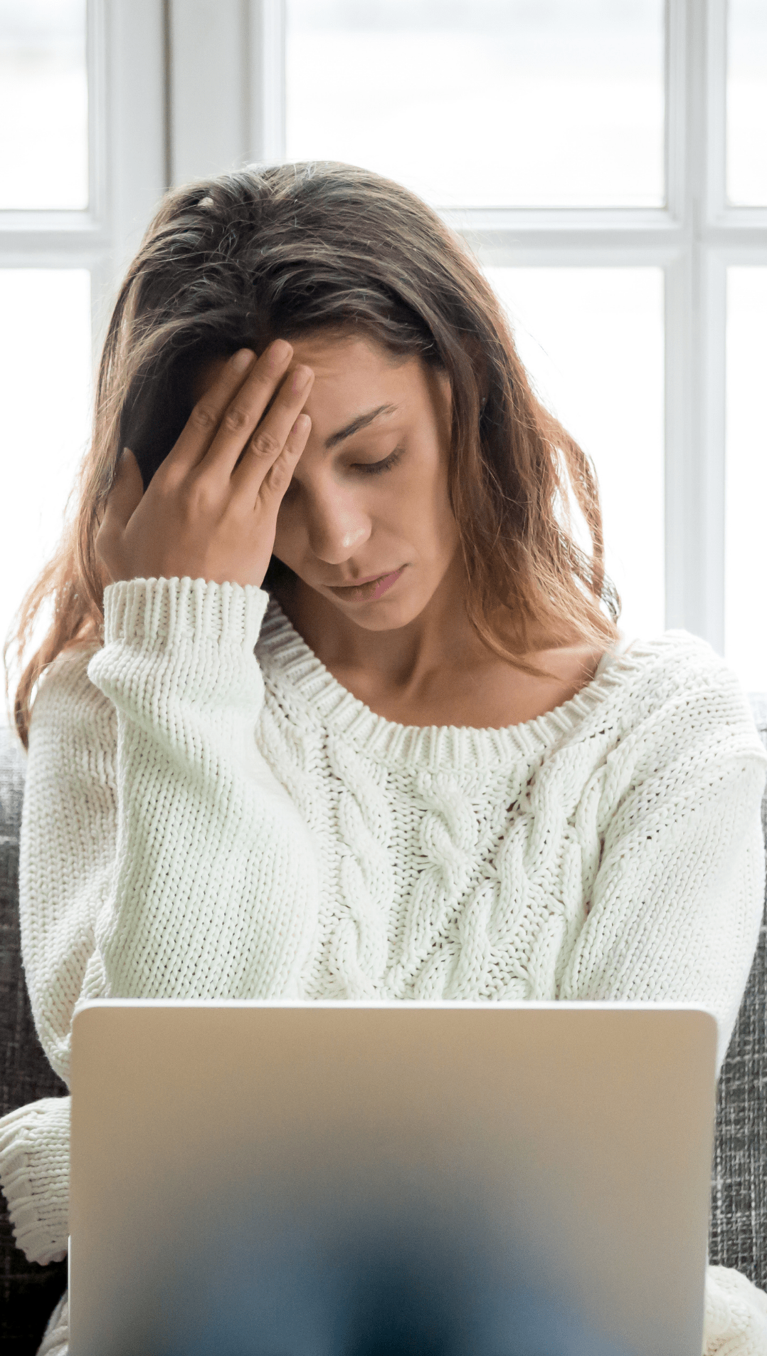 A woman who doesn't feel well is sitting on a couch using a laptop computer.